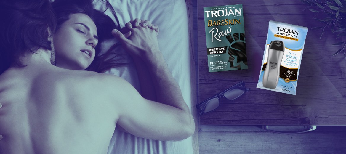 Couple having missionary sex with Trojan condoms and lubricants on bedside table.